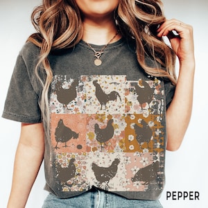 May include: A charcoal gray t-shirt with a vintage-inspired design featuring a rooster and hens in a patchwork pattern with floral accents. The text "PEPPER" is visible in the bottom right corner.