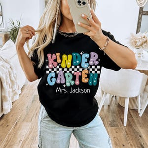 May include: Black t-shirt with the word "KINDERGARTEN" in colorful bubble letters, a checkered pattern, and the name "Mrs. Jackson" below. The shirt is a casual style.