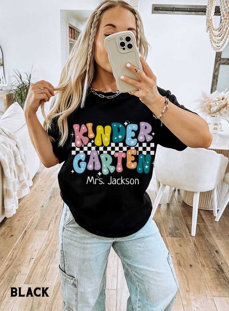 May include: Black t-shirt with the word "KINDERGARTEN" in colorful bubble letters, a checkered pattern, and the name "Mrs. Jackson" below. The shirt is a casual style.
