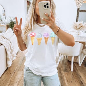May include: White t-shirt with a graphic of four ice cream cones in different colors. The ice cream cones are in a row, with the pink ice cream cone on the left and the blue ice cream cone on the right.