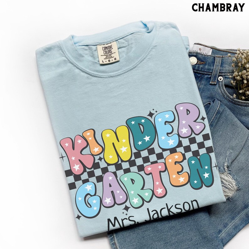 May include: Light blue t-shirt with the word "KINDERGARTEN" in colorful bubble letters with star accents. The shirt has a black and white checkered pattern behind the letters and the name "Mrs. Jackson" below the word.