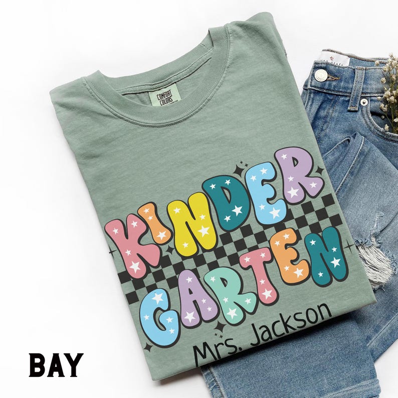 May include: A sage green t-shirt with the word "KINDERGARTEN" in colorful bubble letters with star accents. The shirt also has the text "Mrs. Jackson" and is paired with blue denim jeans.
