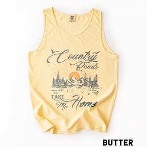 K&ouml;nnte beinhalten: Hellgelbes Tanktop mit dem Text "Country Roads Take Me Home" in verblichener Schrift. Die Grafik zeigt eine Berglandschaft mit aufgehender Sonne und B&auml;umen. Das Tanktop hat ein klassisches, &auml;rmelloses Design.