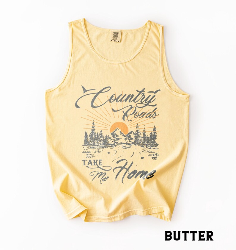 K&ouml;nnte beinhalten: Hellgelbes Tanktop mit dem Text "Country Roads Take Me Home" in verblichener Schrift. Die Grafik zeigt eine Berglandschaft mit aufgehender Sonne und B&auml;umen. Das Tanktop hat ein klassisches, &auml;rmelloses Design.