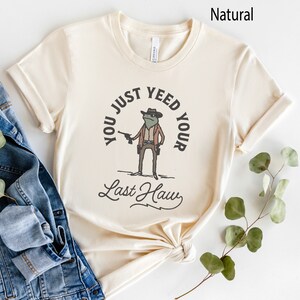 Puede incluir: Una camiseta beige con un dibujo de una rana de dibujos animados con un sombrero de vaquero y sosteniendo una pistola. El texto en la camiseta dice "You just yeed your last haw".