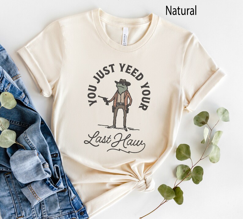 Puede incluir: Una camiseta beige con un dibujo de una rana de dibujos animados con un sombrero de vaquero y sosteniendo una pistola. El texto en la camiseta dice "You just yeed your last haw".