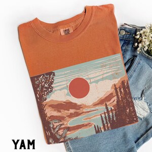 May include: Orange t-shirt with a landscape graphic featuring a red sun, mountains, and a river. The shirt has a crew neck and the text "YAM" at the bottom. The shirt is paired with blue denim jeans.