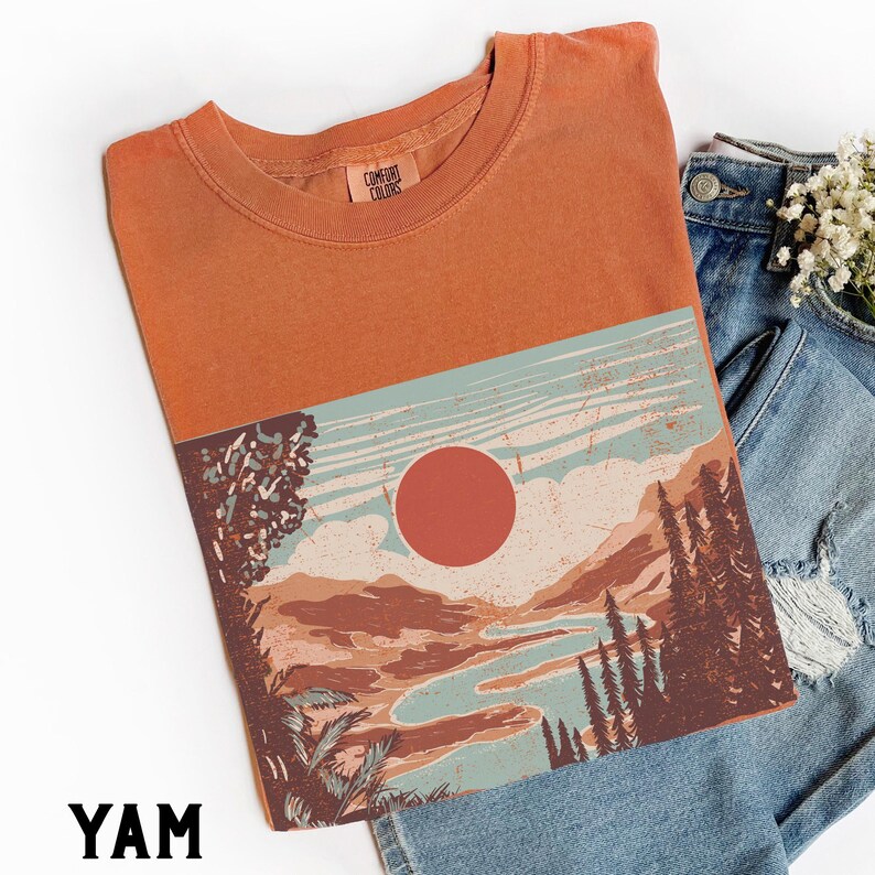 May include: Orange t-shirt with a landscape graphic featuring a red sun, mountains, and a river. The shirt has a crew neck and the text "YAM" at the bottom. The shirt is paired with blue denim jeans.