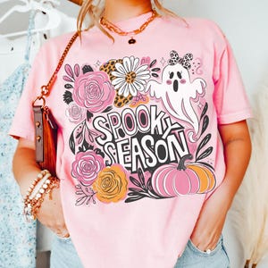 May include: Pink t-shirt with a Halloween-themed graphic. The design includes a ghost, flowers, and a pumpkin, with the words "SPOOK SEASON" in a playful font. The word "BLOSSOM" is printed at the bottom.