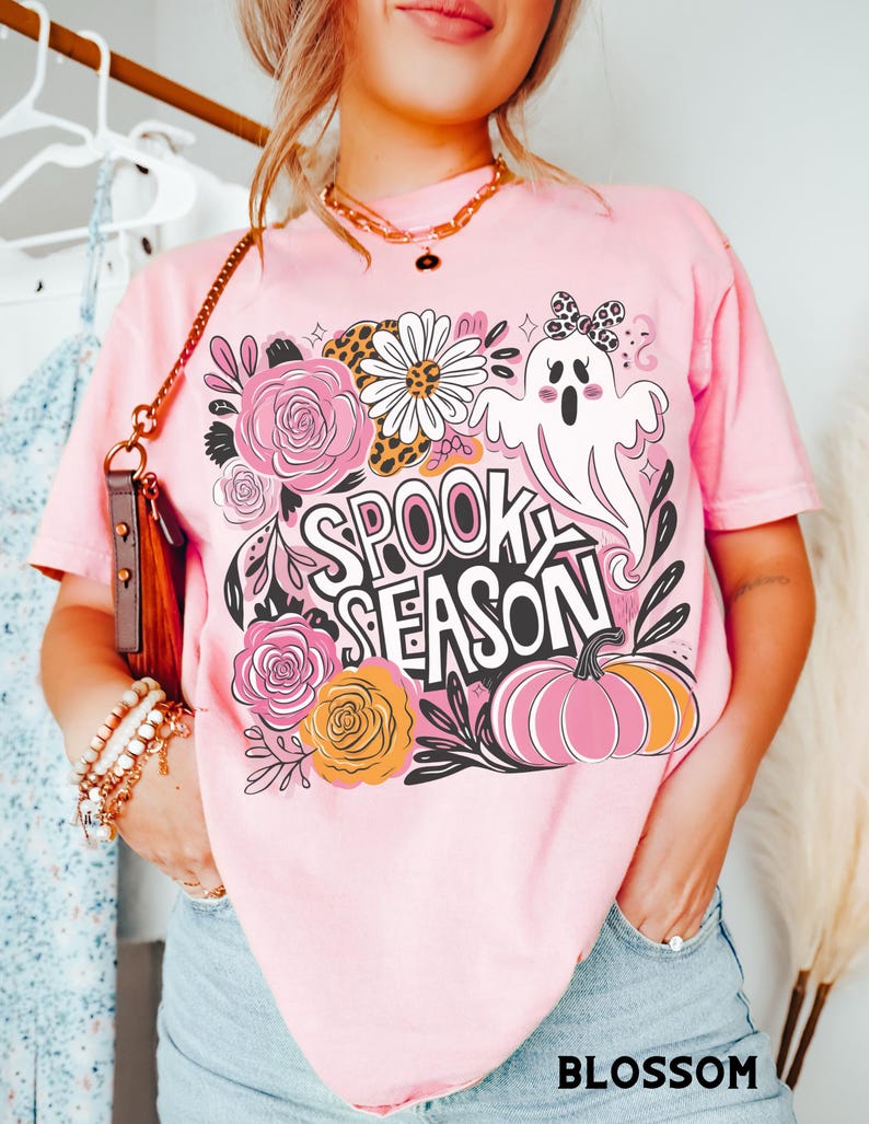 May include: Pink t-shirt with a Halloween-themed graphic. The design includes a ghost, flowers, and a pumpkin, with the words "SPOOK SEASON" in a playful font. The word "BLOSSOM" is printed at the bottom.