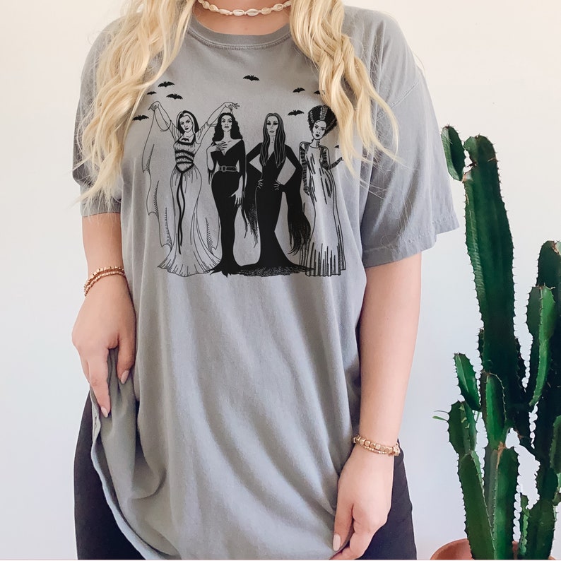 K&ouml;nnte beinhalten: Ein graues T-Shirt mit einem schwarz-wei&szlig;en Grafikdruck von vier Frauen, die als ikonische Horrorfilmfiguren verkleidet sind. Die Frauen stehen in einer Reihe und l&auml;cheln alle. Die Grafik enth&auml;lt Flederm&auml;use, die um die Frauen herumfliegen.