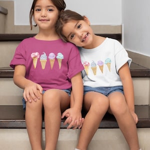 May include: Two young girls wearing matching t-shirts with a graphic of ice cream cones. The girl on the left is wearing a pink shirt and the girl on the right is wearing a white shirt. They are sitting on a set of stairs.