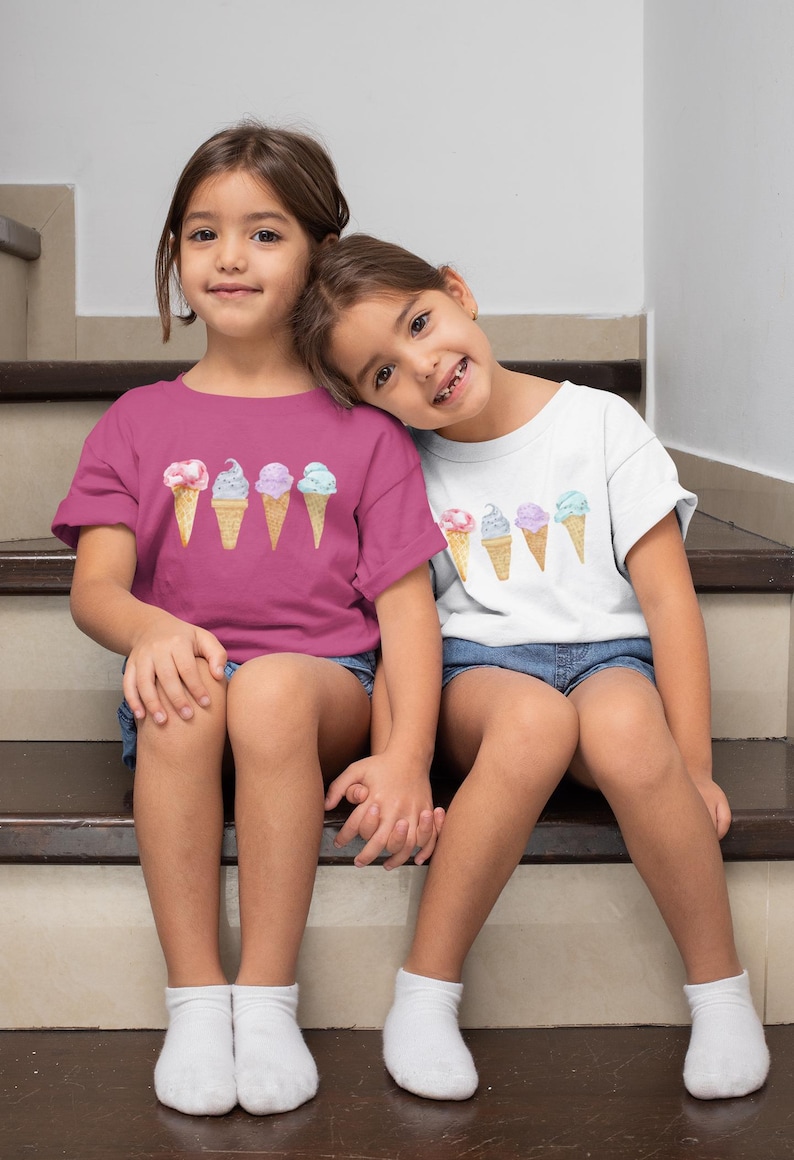 May include: Two young girls wearing matching t-shirts with a graphic of ice cream cones. The girl on the left is wearing a pink shirt and the girl on the right is wearing a white shirt. They are sitting on a set of stairs.