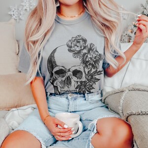 May include: A gray t-shirt featuring a detailed skull design with floral accents. The graphic is in black and white. The word "GRANITE" is printed in black at the bottom of the shirt. The model is wearing denim shorts.