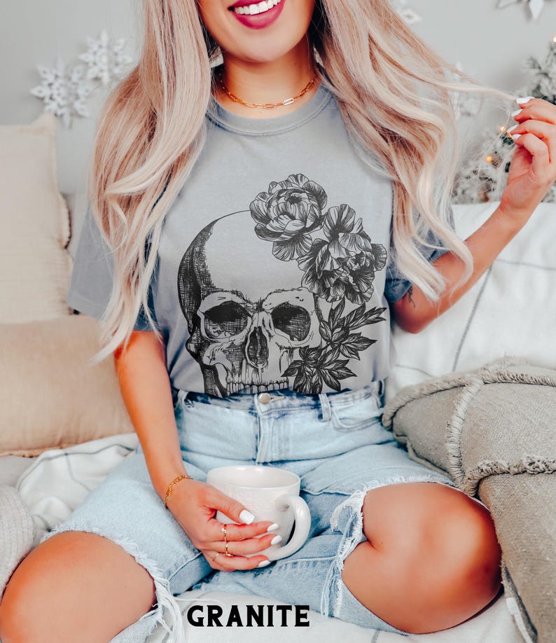 May include: A gray t-shirt featuring a detailed skull design with floral accents. The graphic is in black and white. The word "GRANITE" is printed in black at the bottom of the shirt. The model is wearing denim shorts.