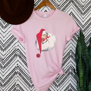 May include: A pink t-shirt with a vintage illustration of Santa Claus on the front. The illustration is in red, white, and black.