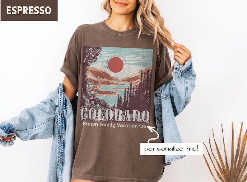 May include: Espresso-colored t-shirt featuring a Colorado landscape design with a red sun, mountains, and a lake. The text "COLORADO" and "Brown Family Vacation '26" are printed on the front. The model is wearing a denim jacket.