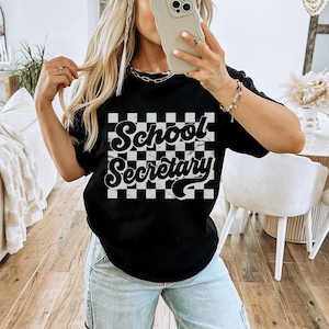 Peut inclure: T-shirt noir avec un fond &agrave; carreaux blancs et le texte "School Secretary" dans une police r&eacute;tro.
