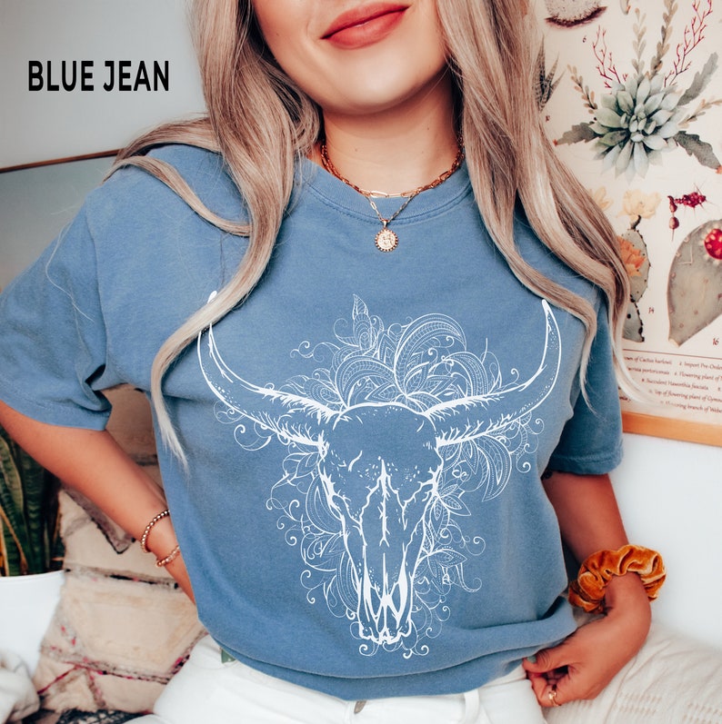 K&ouml;nnte beinhalten: Ein blaues Jeans-T-Shirt mit einem wei&szlig;en Grafikdruck eines Bullensch&auml;dels, umgeben von floralen Designs. Der Text "BLUE JEAN" ist in der oberen linken Ecke des Bildes sichtbar.