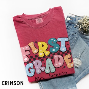 May include: Crimson-colored t-shirt with the words "FIRST GRADE" in colorful bubble letters, a black and white checkered pattern, and the name "Mrs. Jackson". The shirt is paired with denim shorts.