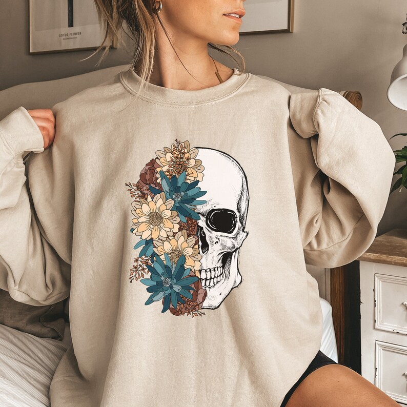 May include: A beige sweatshirt with a graphic of a skull with flowers covering one side of the skull. The flowers are in shades of blue, yellow, and brown.