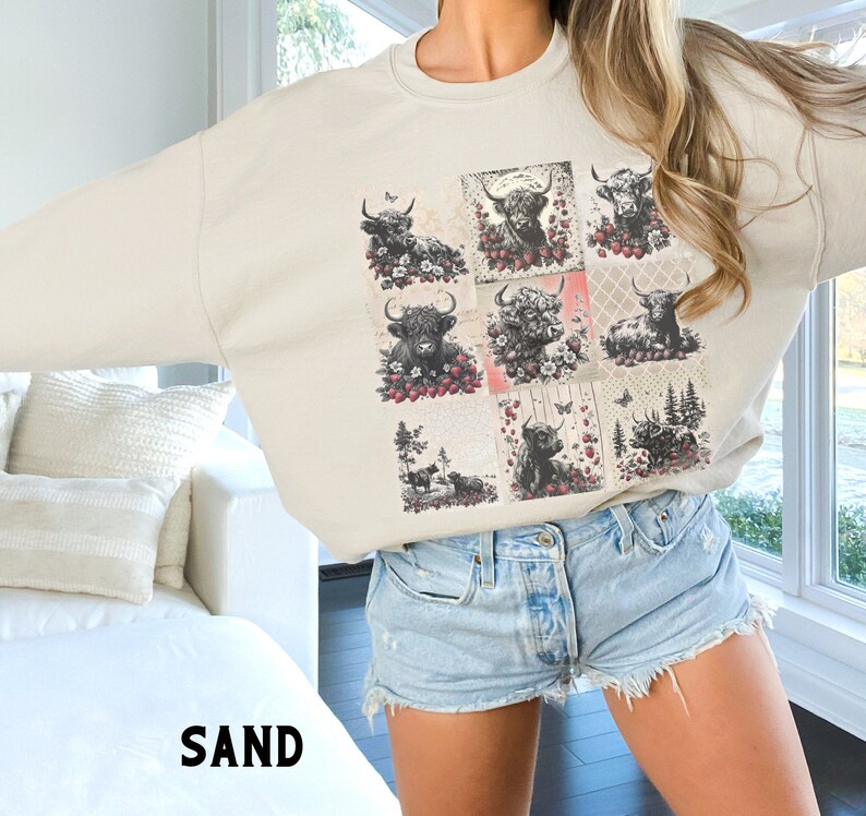 May include: A beige sweatshirt with a graphic print of a patchwork design featuring multiple images of Highland cattle with flowers and butterflies. The sweatshirt is being worn by a person wearing denim shorts.