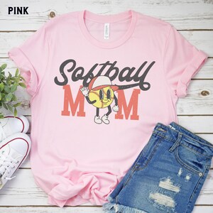 May include: Pink t-shirt with the words "Softball Mom" in a retro font. The design includes a cartoon baseball character wearing a hat. The shirt is paired with denim shorts and white sneakers.