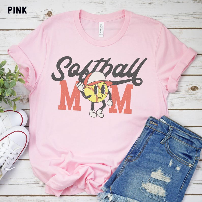 May include: Pink t-shirt with the words "Softball Mom" in a retro font. The design includes a cartoon baseball character wearing a hat. The shirt is paired with denim shorts and white sneakers.