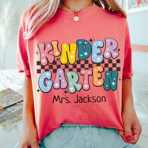 May include: A coral-colored t-shirt with the words "KINDERGARTEN" in colorful, bubble letters with star accents. Below the text is "Mrs. Jackson" in black. The shirt has a checkered pattern and the word "WATERMELON" at the bottom.