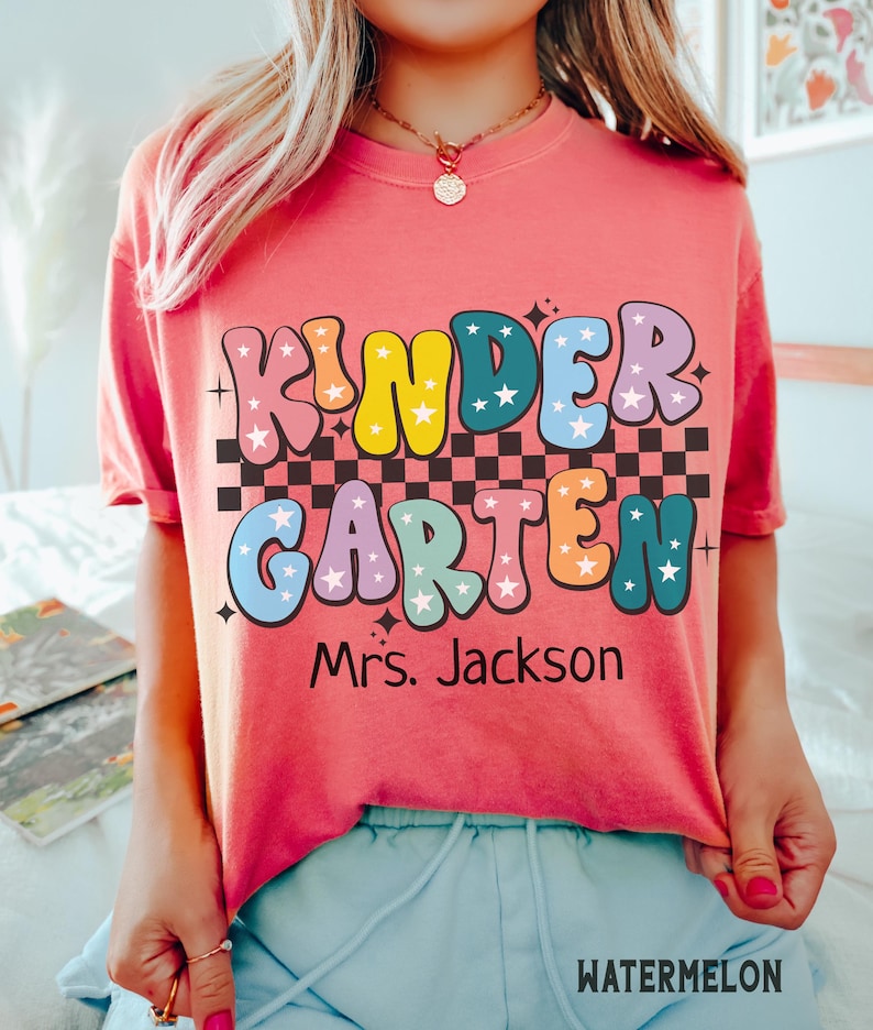 May include: A coral-colored t-shirt with the words "KINDERGARTEN" in colorful, bubble letters with star accents. Below the text is "Mrs. Jackson" in black. The shirt has a checkered pattern and the word "WATERMELON" at the bottom.
