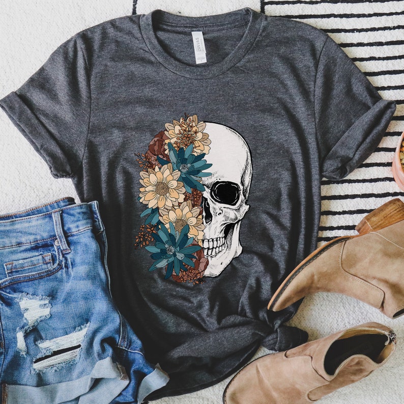 Op de afbeelding: Een donkergrijs T-shirt met een wit schedelontwerp. De schedel is gedeeltelijk bedekt met een boeket kleurrijke bloemen, waaronder blauwe, gele en oranje.