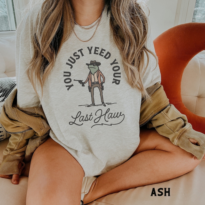 Puede incluir: Una sudadera gris claro con un gr&aacute;fico de una rana con un sombrero de vaquero y sosteniendo una pistola. El texto "You Just Yee'd Your Last Haw" est&aacute; impreso en la sudadera.