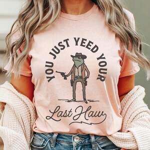 Puede incluir: Una camiseta rosa con un gr&aacute;fico de una rana con un sombrero de vaquero y sosteniendo una pistola. El texto en la camiseta dice "You Just Yee'd Your Last Haw".