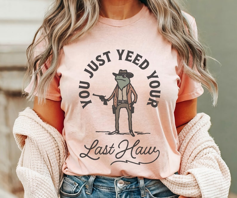 Puede incluir: Una camiseta rosa con un gr&aacute;fico de una rana con un sombrero de vaquero y sosteniendo una pistola. El texto en la camiseta dice "You Just Yee'd Your Last Haw".