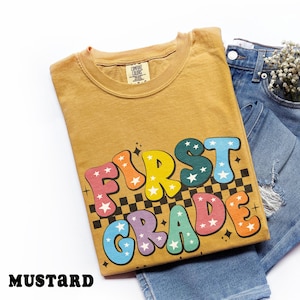 May include: Mustard-colored t-shirt with the words "FIRST GRADE" in colorful, bubble letters with star accents. The shirt is folded and placed on top of a pair of blue denim jeans.