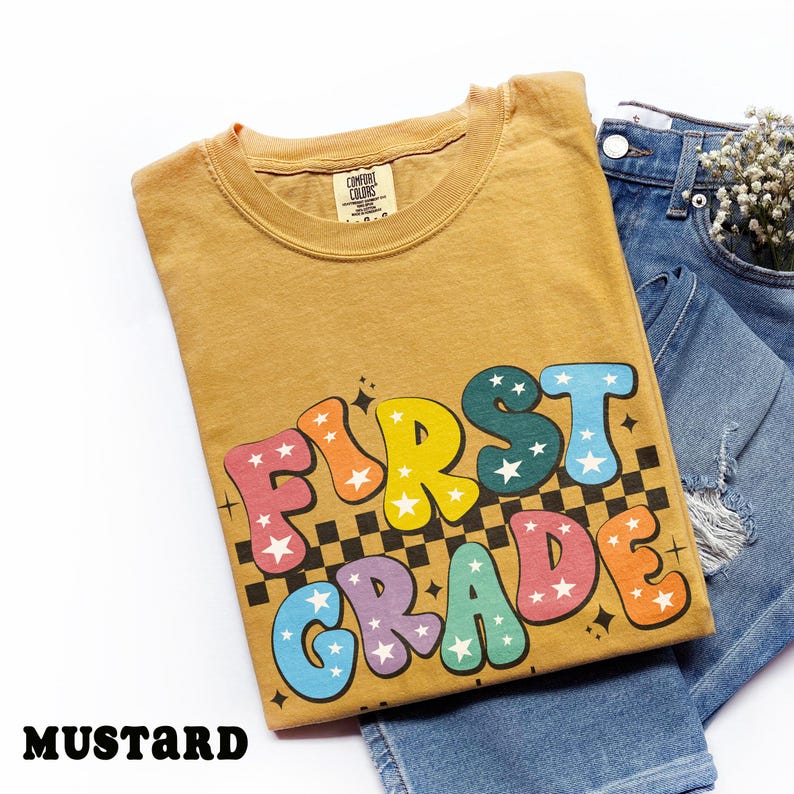 May include: Mustard-colored t-shirt with the words "FIRST GRADE" in colorful, bubble letters with star accents. The shirt is folded and placed on top of a pair of blue denim jeans.