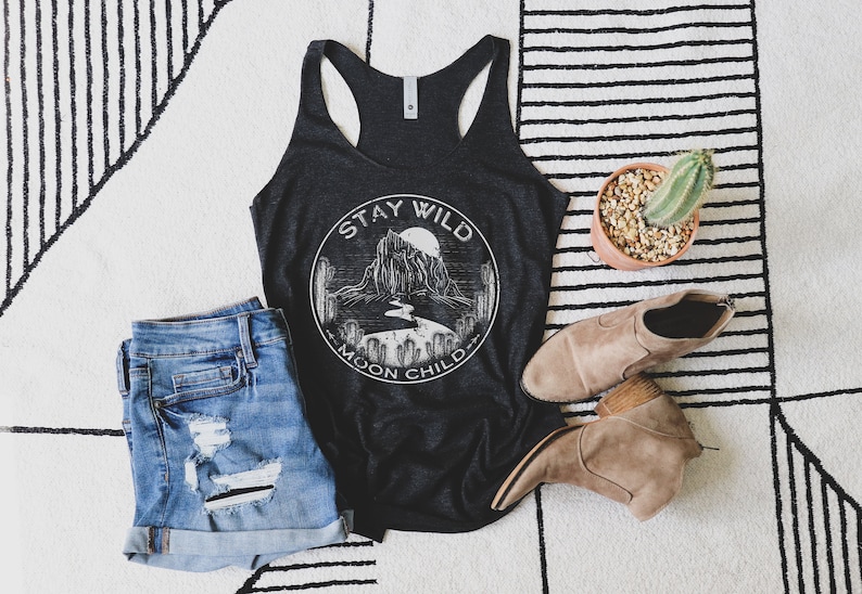 May include: A black tank top with a white graphic of a mountain range and the text "Stay Wild Moon Child". The tank top is laying on a white and black striped rug with a pair of brown ankle boots and a potted cactus.