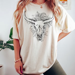 K&ouml;nnte beinhalten: Ein cremefarbenes T-Shirt mit einem schwarz-wei&szlig;en Grafikdruck eines Bullensch&auml;dels mit floralen Details.