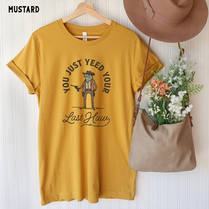 Puede incluir: Una camiseta amarilla mostaza con un dibujo de una rana de dibujos animados con un sombrero de vaquero y sosteniendo una pistola. El texto en la camiseta dice "You Just Yeed Your Last Haw".