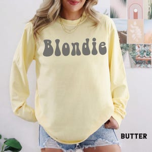 Puede incluir: Una camiseta de manga larga de color amarillo p&aacute;lido con la palabra "Blondie" en letras grises en la parte delantera. La camiseta es de color liso y tiene cuello redondo. La palabra "Butter" est&aacute; impresa en blanco en la parte inferior derecha.
