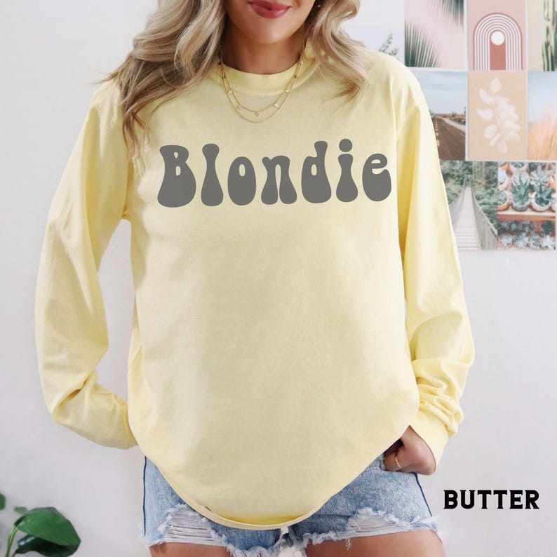 Puede incluir: Una camiseta de manga larga de color amarillo p&aacute;lido con la palabra "Blondie" en letras grises en la parte delantera. La camiseta es de color liso y tiene cuello redondo. La palabra "Butter" est&aacute; impresa en blanco en la parte inferior derecha.