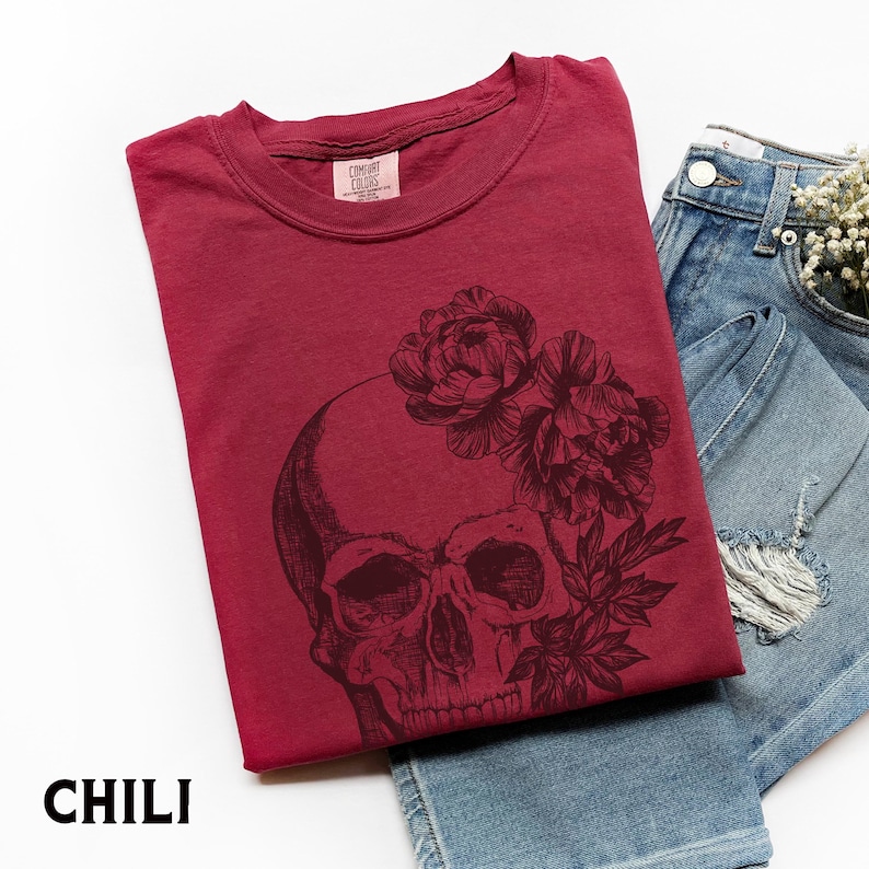 May include: A maroon t-shirt with a black skull and floral design. The shirt is folded next to a pair of blue denim shorts. The word "CHILI" is printed at the bottom of the image. The shirt has a crew neck and short sleeves.
