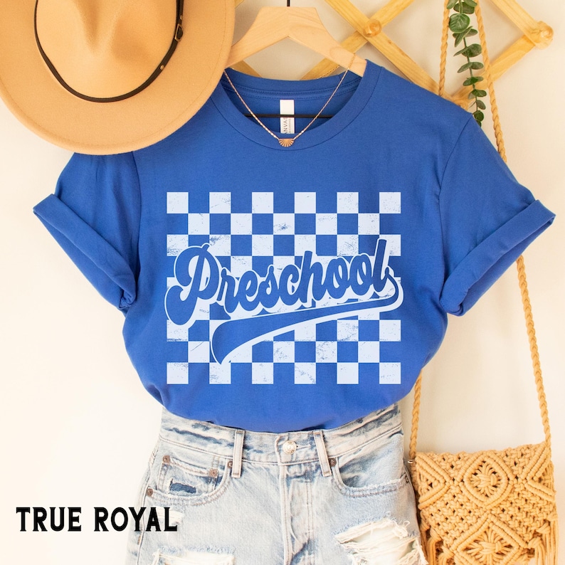 Pu&ograve; includere: Una maglietta blu reale con maniche arrotolate. La maglietta presenta un motivo a scacchi blu chiaro e bianco con la parola "Preschool" in corsivo. Il testo "TRUE ROYAL" &egrave; in basso.