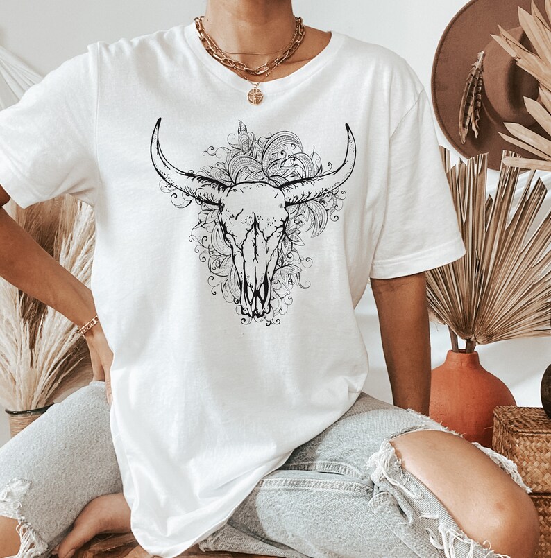 Peut inclure: T-shirt blanc avec un motif noir et blanc de cr&acirc;ne de taureau avec des d&eacute;tails floraux.