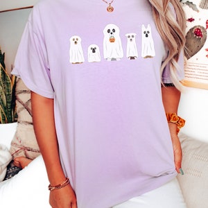 May include: A lavender t-shirt with a graphic of five ghost dogs. The dogs are wearing ghost sheets and have different expressions. One dog is holding a small orange pumpkin bucket.