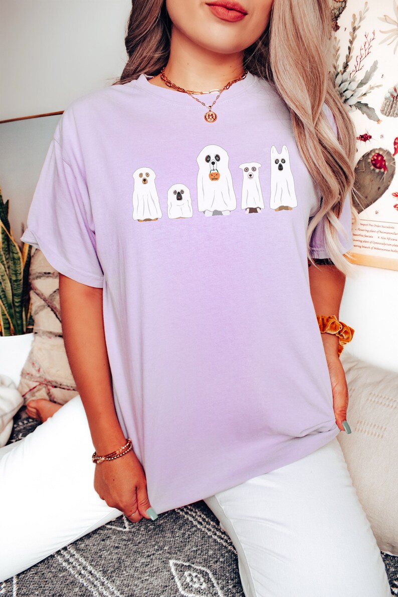 May include: A lavender t-shirt with a graphic of five ghost dogs. The dogs are wearing ghost sheets and have different expressions. One dog is holding a small orange pumpkin bucket.