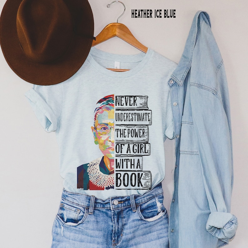Op de afbeelding: Lichtblauw T-shirt met een afbeelding van een vrouwengezicht en de tekst "NEVER UNDERESTIMATE THE POWER OF A GIRL WITH A BOOK." Het shirt hangt aan een houten hanger. De woorden "HEATHER ICE BLUE" staan bovenaan.