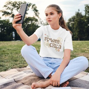 Puede incluir: Camiseta blanca con el texto "FLOWER CHILD" en marr&oacute;n y beige, con detalles florales. La persona que lleva la camiseta est&aacute; sentada en una manta en una zona de c&eacute;sped y sostiene un smartphone.