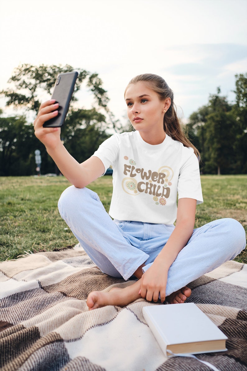 Puede incluir: Camiseta blanca con el texto "FLOWER CHILD" en marr&oacute;n y beige, con detalles florales. La persona que lleva la camiseta est&aacute; sentada en una manta en una zona de c&eacute;sped y sostiene un smartphone.