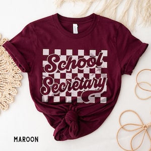 Peut inclure: T-shirt marron avec un fond &agrave; carreaux blancs et le texte "School Secretary" dans une police r&eacute;tro.